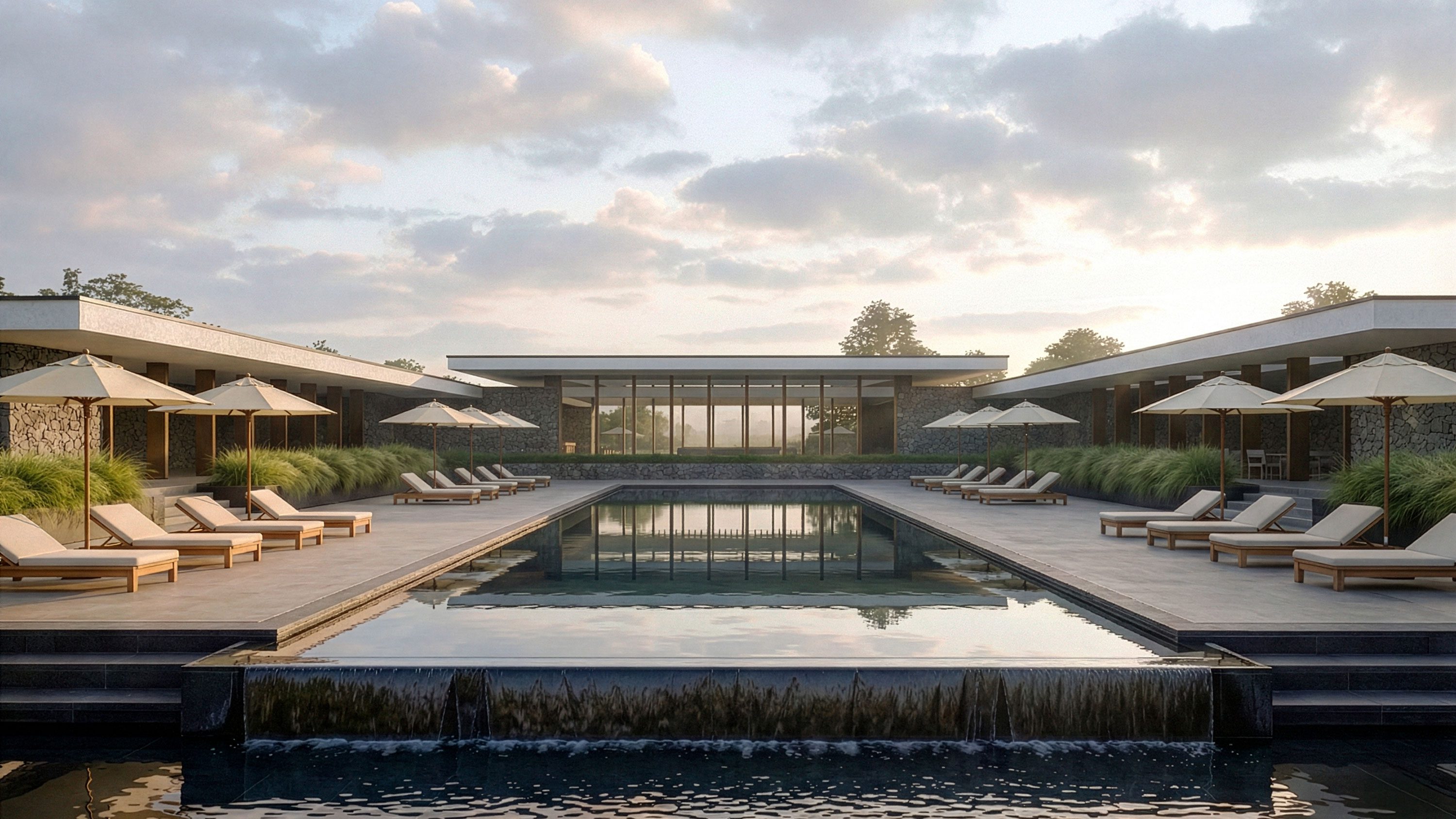 Symmetrical hotel pool courtyard with infinity edge, lounge chairs, umbrellas, native grasses, and stone-clad wings at dusk.