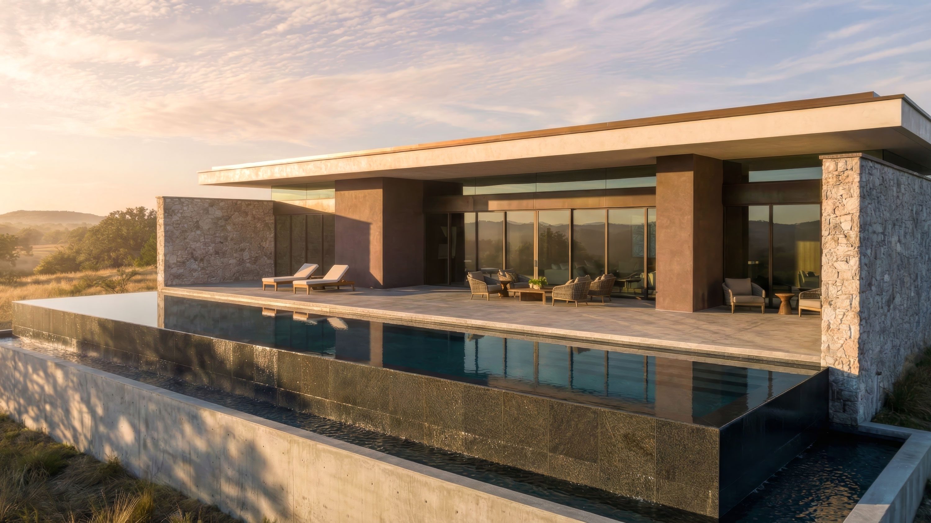 Modern home rear exterior with infinity pool, stone and stucco facade, glass walls, and rolling hill views at sunset.