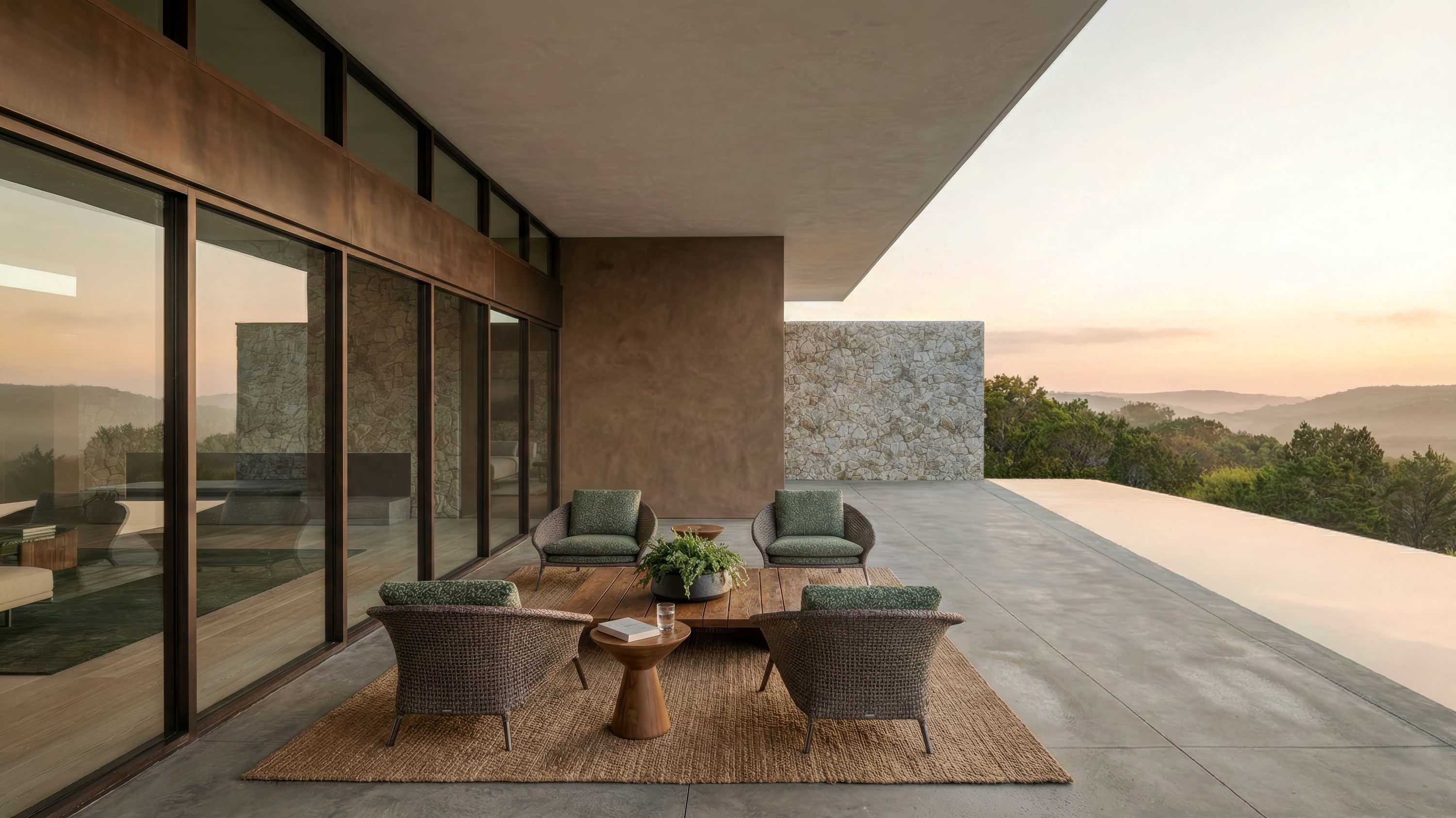Covered terrace with woven lounge chairs, wood coffee table, stone accent wall, and panoramic hill country views at dusk.